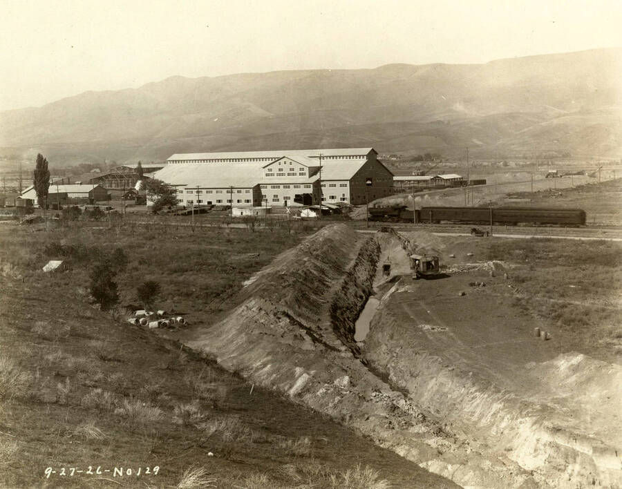 In the foreground, a crane works to bring up dirt. In the middle of the photograph a train is moving past the Lewiston Mill which sits in the background. Written on the photograph is '9/27/1926 No. 129.'