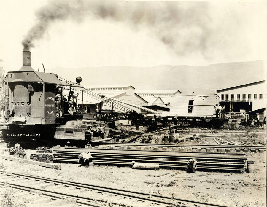 Two different cranes work at the Lewiston Mill to help with construction. The crane in the background has 'Clearwater Timber Co' on it's side while the crane in the foreground has 'American Locomotive Crane' written on it. Written on the photograph as well is '7/16/1927 No. 299.'