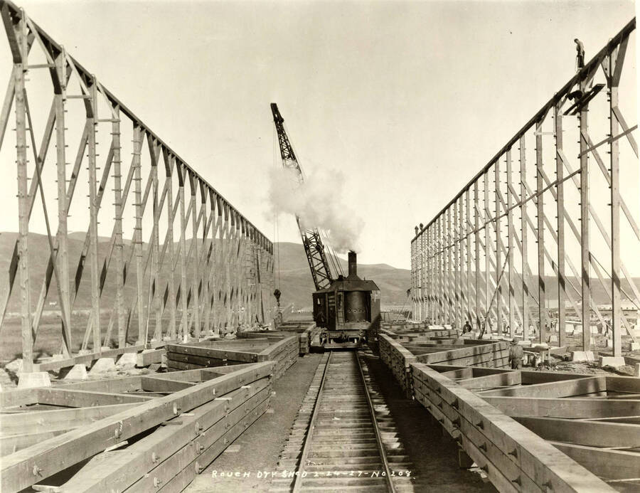 A locomotive crane works to help construct the rough-dry shed at the Lewiston Mill. On the right hand side of the photo a man stands atop the framing. Written on the photograph is 'Rough dry shed, 2/24/1927, No. 208.'