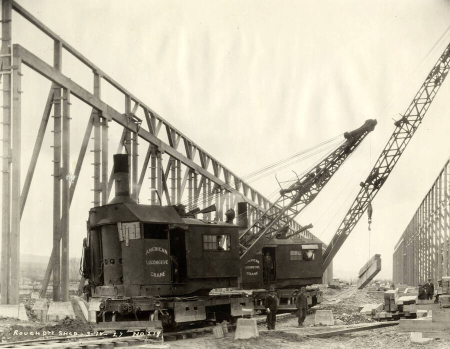 Two locomotive cranes work to help complete construction on the rough-dry shed at the Lewiston Mill. The one in front is lifting bundles of lumber. Both have written on them 'American Locomotive Crane'. Written on the photograph is 'Rough dry shed, 3/18/1927, No. 218.'