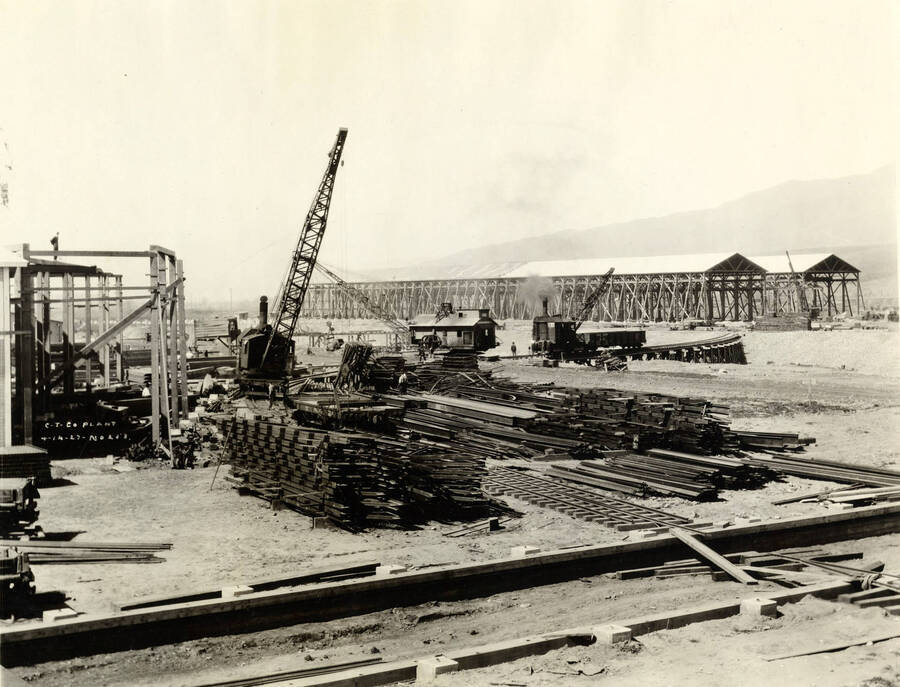 Piles of lumber are stacked waiting to be used for construction. Several cranes work in the background. Also in the background are two half-completed buildings. Written on the photograph is 'CT CO Plant 4/14/1927 No. 203.'