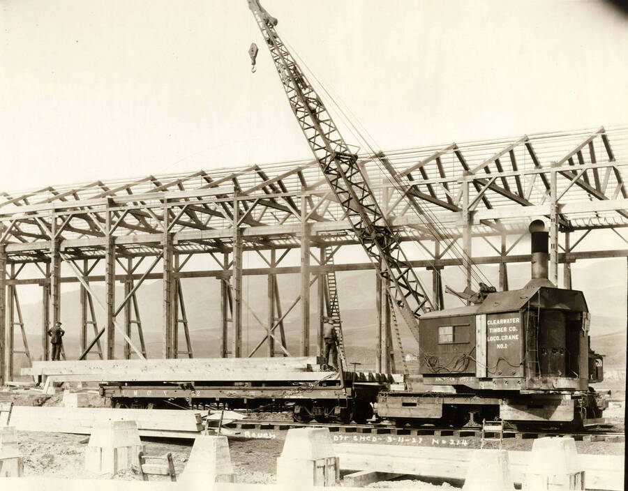 A locomotive crane works to help construct the rough-dry shed at the Lewiston Mill. Two men stand on the flatcar holding the lumber. Written on the photograph is 'Rough dry shed, 3/11/1927, No. 214.'