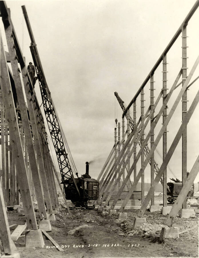 A locomotive crane helps in the construction of the rough-dry shed at the Lewiston Mill. Men work at the top of the frame on the left hand side of the photograph. Written on the photograph is 'Rough dry shed, 3/18/1927, No. 222.'