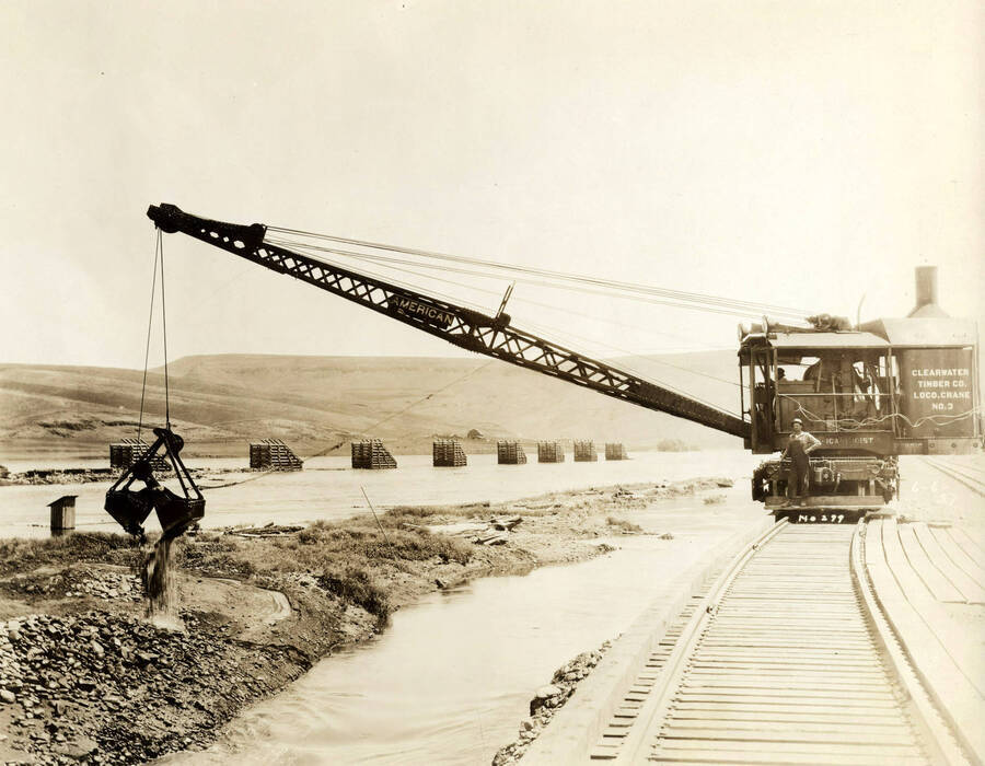 A locomotive crane dumps a bucket full of rocks. The side of the crane reads 'Clearwater Timber Co. Loco. Crane No. 3.'