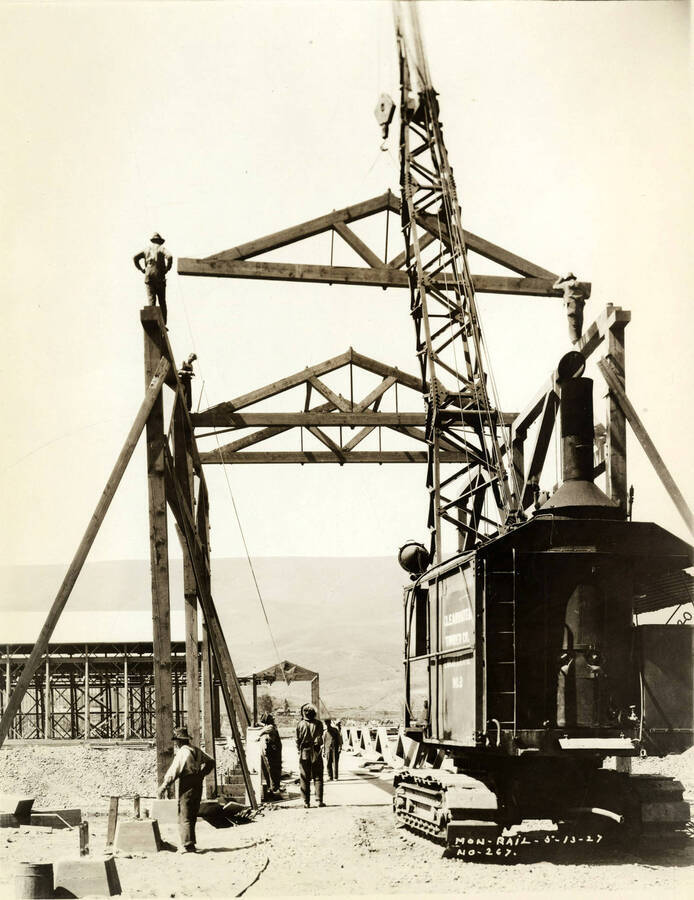A crane lifts a roof truss in the construction of the monorail at the Lewiston Mill. Men stand on either side of the framing to help with construction. Men also stand watching on the ground. Written on the photograph is 'Mon-Rail 5/13/1927 No. 267.'