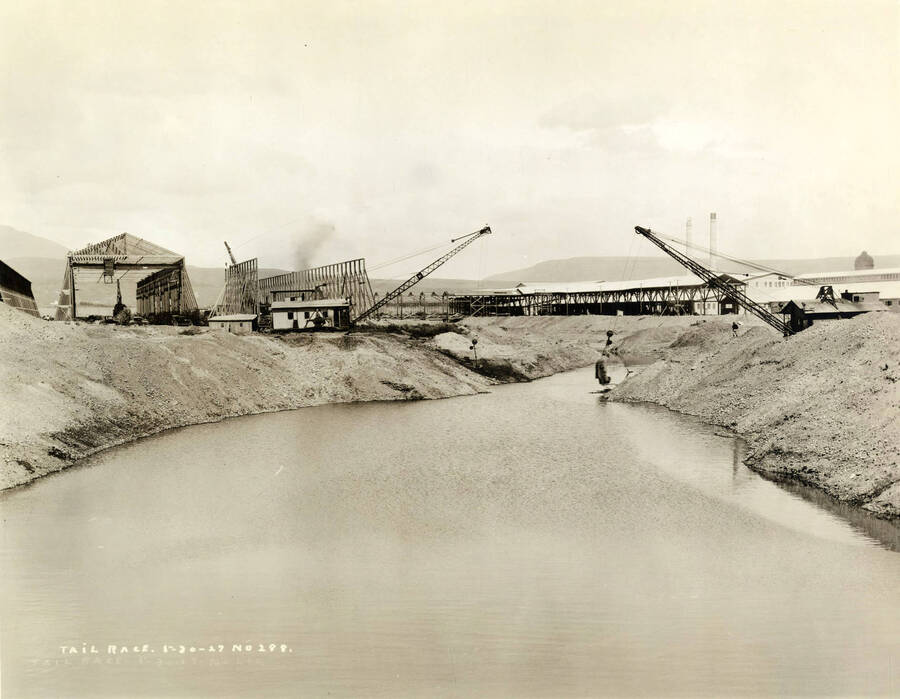 Two cranes work to build the tailrace for the Lewiston Mill. On the left hand side, are two unfinished buildings. Written on the photograph is 'Tail Race, 5/30/1927, No. 288.'