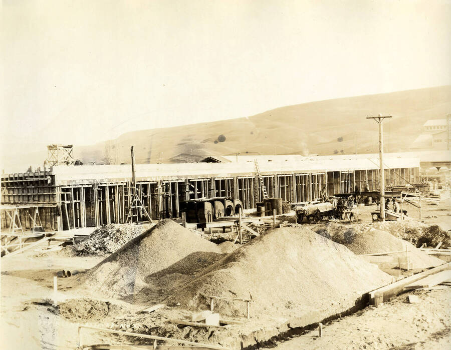 Piles of material lie in front of buildings being constructed. According to the description written on the back of the photograph, these are the dry kilns ('Dry Kilns 2/11/1927 No. X').