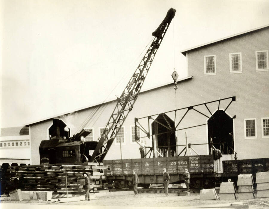 A crane lifts a metal structure of a flatcar to aid in construction of the Lewiston Mill. Four men stand on the ground while another stands on the flatcar.