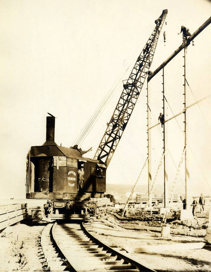 A locomotive crane assisting in building the Rough-Dry shed at the Lewiston Mill. Three men stand at the bottom of the frame, while one man sits halfway up and two stand atop the frame. Written on the photograph is 'Rough Dry shed, 2/24/1927, No. 207.'