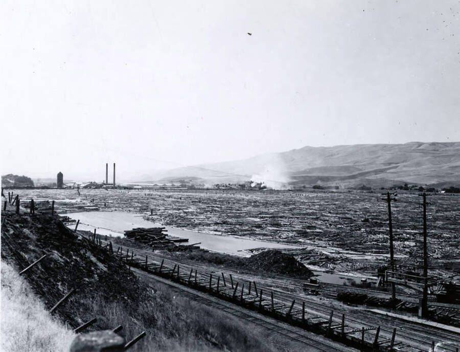 Looking at the log pond from the hillside. At the bottom of the picture sit empty flatcars. In the distance is the sawmill. Note on the envelope says that this photo was taken for Doug Wilson, the Seattle AP photographer who covered the log drive.