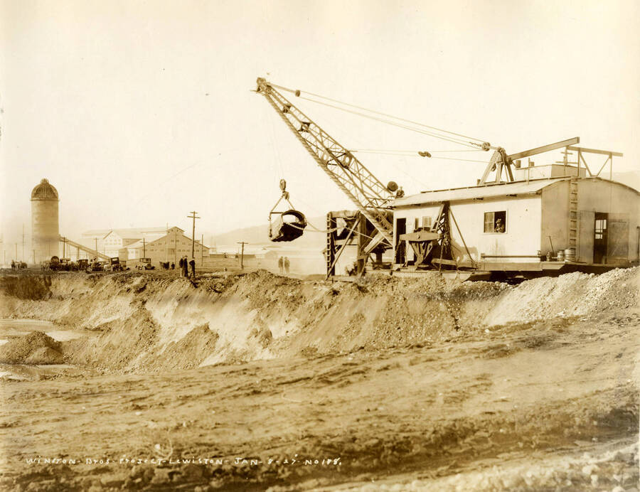 A crane lifts a piece of building material. Two men stand watching. Written on the photograph is 'Winston Brothers Project, 1/8/1927 No. 178.'