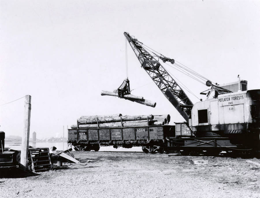 A crane lifts a log into the log pond from a railcar. The description on the back reads "unloading at forebay." Note on the envelope says that this photo was taken for Doug Wilson, the Seattle AP photographer who covered the log drive.