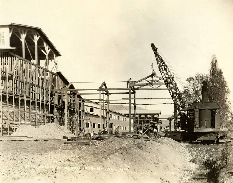 A crane works to help build the roof supports. A man sits atop the roof support near the crane  while another stands on a different part of the framing. Several men work below. Written on the picture is 'Stacker Building and Mon-Rail 10/18/1926 No. 146.'