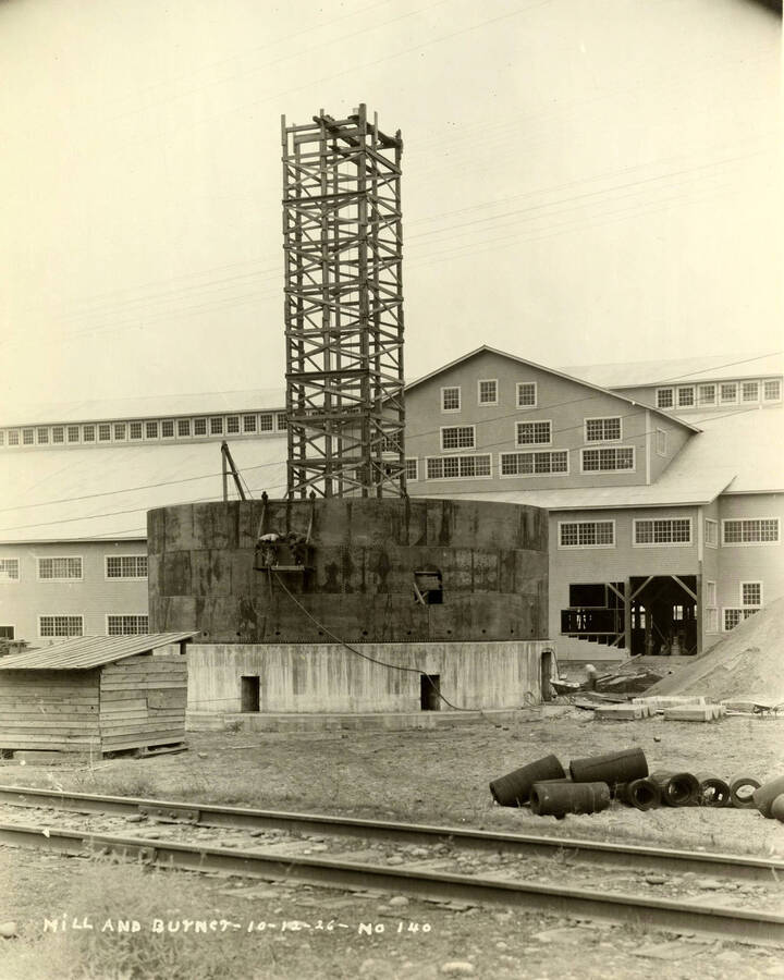 The partially completed burner stands in front of the almost completed mill. Railroad tracks run across the bottom of the photograph. Written on the photograph is 'Mill and burner 10/12/1926 No. 140'