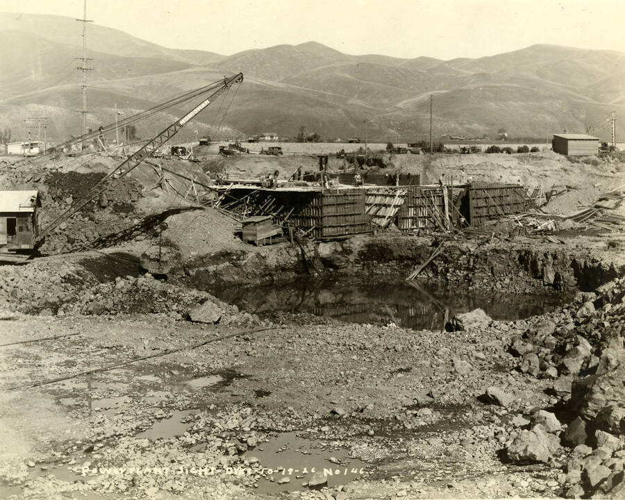 A crane lifts dirt of a pool of water. In the background, men work on the power plant. Written on the photograph is 'Power Plant site with Dike 10/19/1926 No. 146'