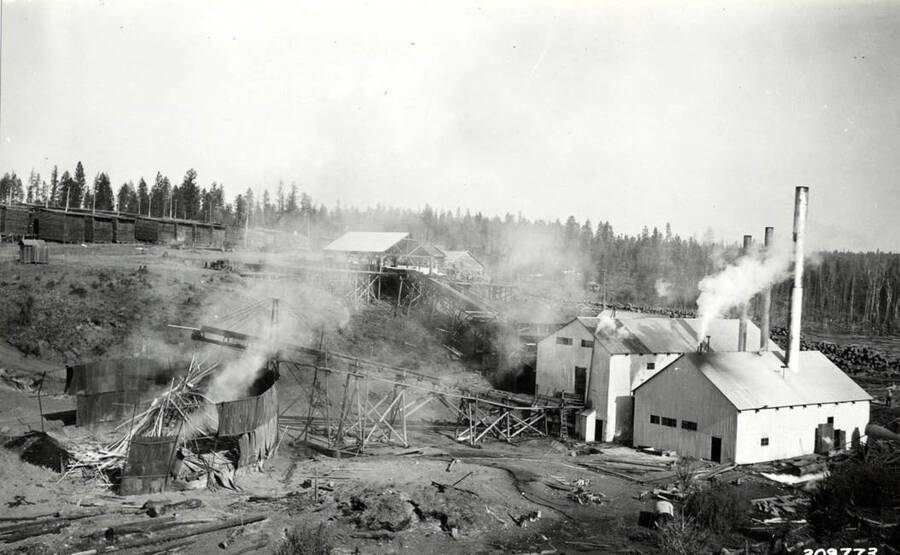 Part of the one of the Potlatch mills (not Lewiston). On the left hand side, a structure appears to be smoldering due to fire. On the right sit mill buildings . In the background on the left side of the picture is the lumber yards.