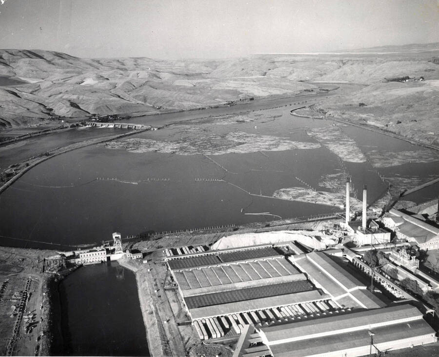 An aerial photograph of the Clearwater Paper Mill and log pond. Clearwater dam is in the upper left hand side of the photograph. On the back of the photograph, a stamp says "Please credit when used the Washington Water Power CO. Spokane, Washington." It lists John Keith as the photographer.