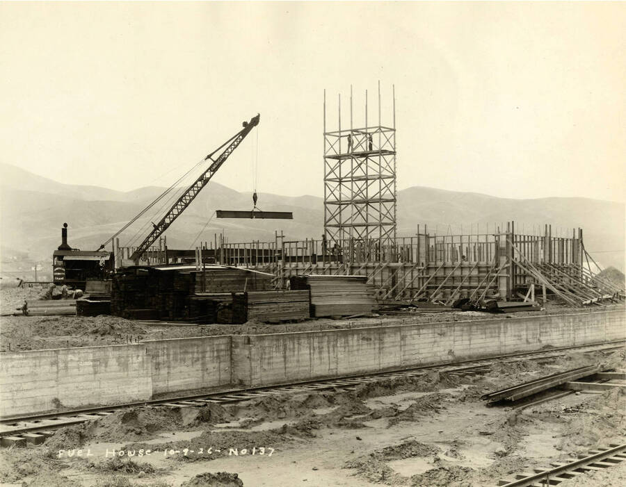 A crane lifts a steel beam on the  construction of the fuel house. Two men stand atop a wooden scaffold. Written on the picture is 'Fuel house.  10/9/1926 No. 137'
