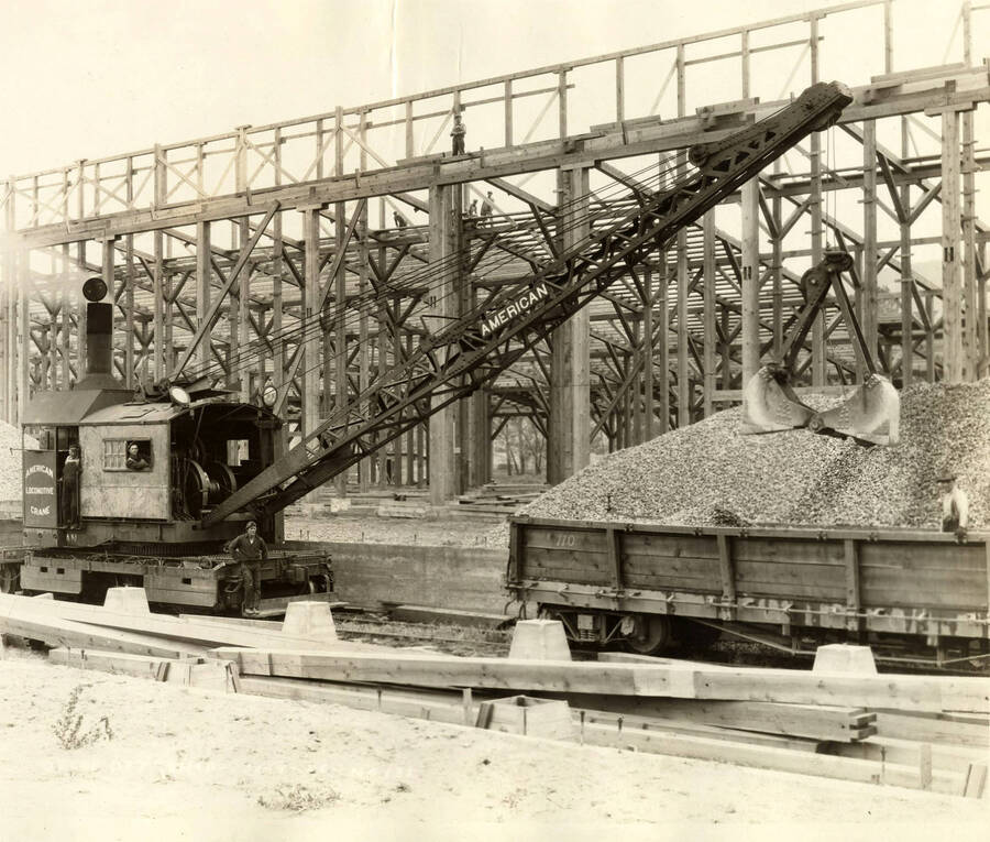 A crane dumps gravel into a railcar. Behind the crane is the framing for one of the drying sheds. Written on the photograph is 'Drest Dry shed, 10/9/1926, No. 135'