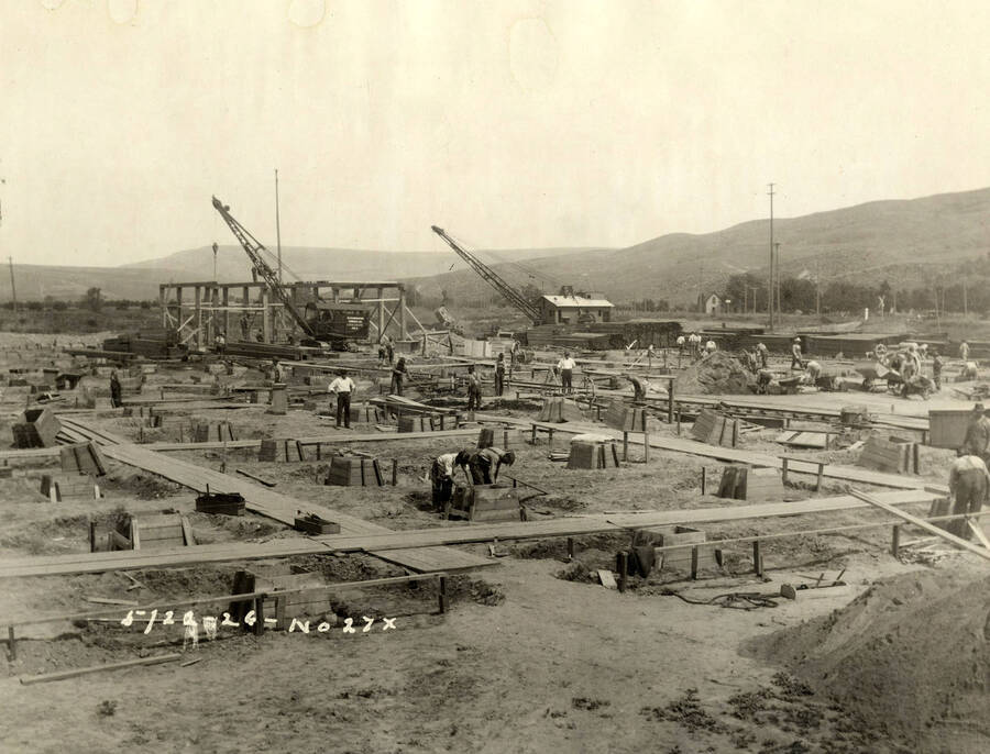 Construction of one of the buildings at the Lewiston Mill. In the center of the photograph, two men work on a cement form. In the background are two cranes. Written on the photograph is '5/22/1926 No. 27x'