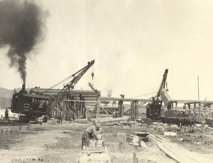 Two cranes lift loads of lumber to help build one of the buildings at the Lewiston Mill. A man works in the foreground on one of the foundation frames.  Written on the photograph is '5/25/1926 No. 34'