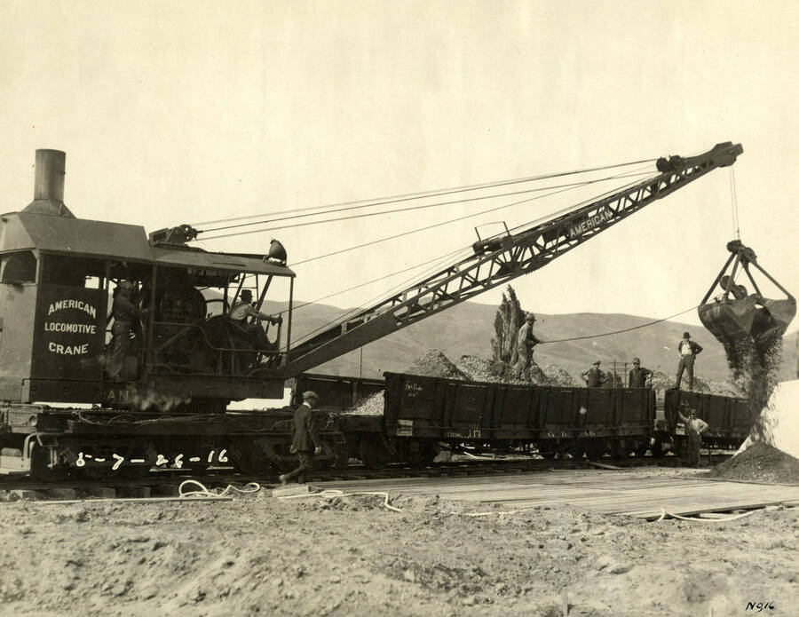 A locomotive crane dumps gravel and dirt onto boards. A man in a suit walks toward the pile of dirt and gravel while other men look on. The wording on the locomotive crane reads 'American Locomotive Crane.' Written on the photograph is '5/7/1926 No. 16'