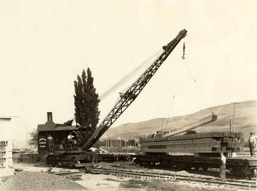 A locomotive crane picks up a piece of lumber. One man stands on the flatcar while another next to it. Three men stand either on the crane or next to it. Written on the photograph is '5/22/1926 No. 30'