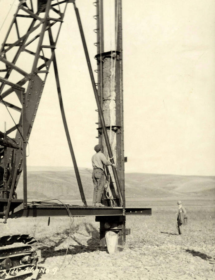 A pile driver pushes a piling into the ground. One man stands on crane holding the pile driver while another man stands in the background. Written on the photograph is 'The first piling. 2/15/1926 No. 9'