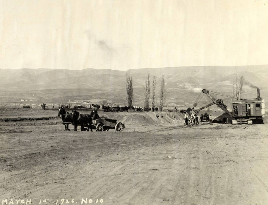 In the foreground, horse-drawn wagon wheels away from a crane dumping into another wagon in the background. Also in the background, stand horses, men, and horses hitched to wagons. Written on the photograph is '3/15/1926 No. 10'