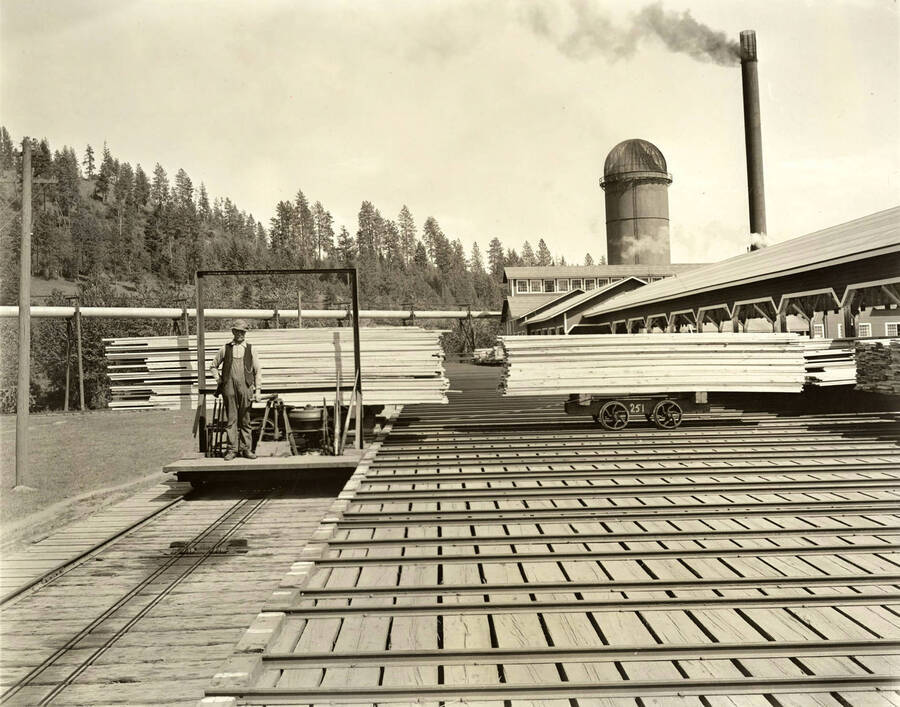 A man steers a flatcar full of lumber while another flatcar on his left waits. In the background are buildings of the Rutledge Mill in Coeur d'Alene, Idaho. ON the back of the photograph it says 'Rutledge Mill'