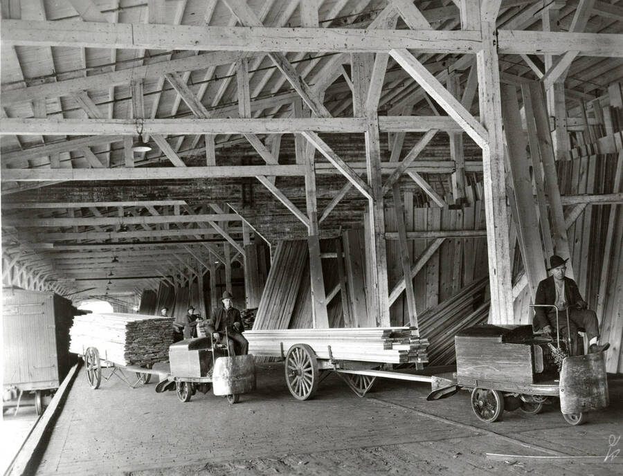Two men sit on carts pulling trailers full of finished lumber boards. Two more men stand in the background. To the right of all the men are different holding areas full of lumber.