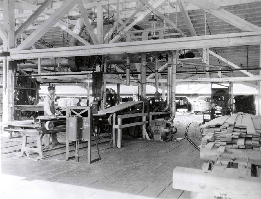 A man works on a piece of woodworking machinery. Written on the back of the photograph is "Edward Rutledge Timber Co. - Coeur d'Alene, Idaho. Machinery in remanufacturing building looking towards the planing mill."