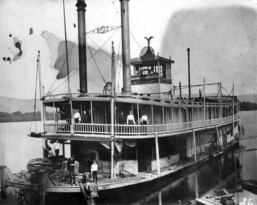 The steamboat owned by the Edward Rutledge Timber Company. On the first deck men stand waiting as two men haul up a wheelbarrow. Five women and a young child stand on the second deck watching.