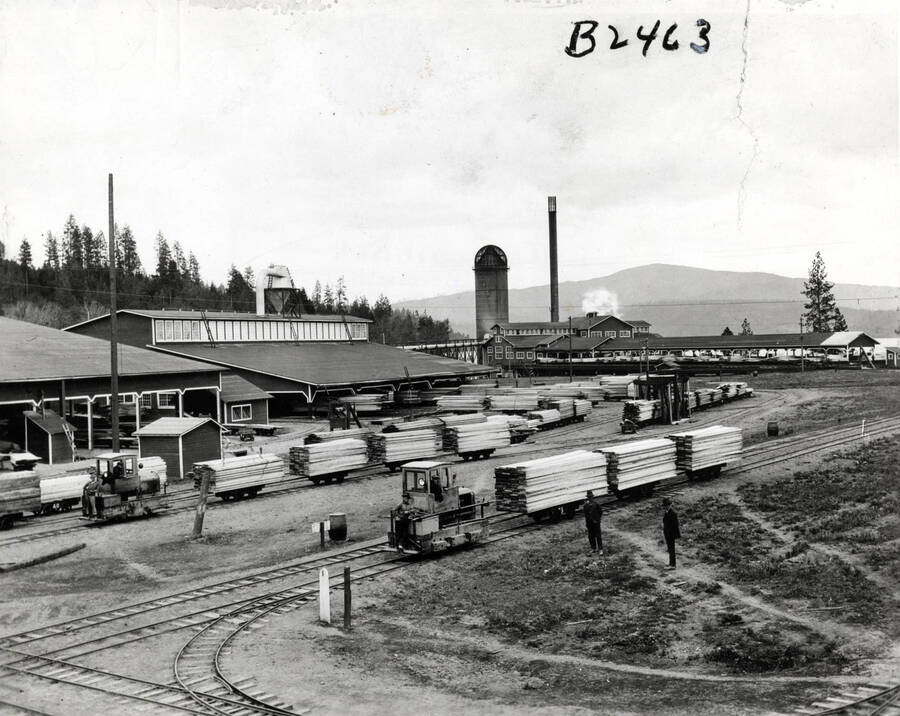A view of the Rutledge Mill lumber Mill. Two locomotive systems pull flatcars full of lumber. In the distance, Lake Coeur d'Alene can been seen. Stamped on the back is the stamp of the photographer, John Bickel with is Tacoma, Washington address.