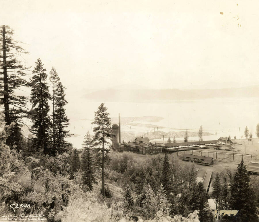 Looking down from the hill behind the mill. In view are the mill, saw mill, the log ponds, and Lake Coeur d'Alene. On the photograph in the bottom left-hand corner is 'John D. Cress 'Forest Fotografer' 1330 7th, Cor. Union, Seattle Wash. The description on the back reads 'Two section panorama of the plant from hill. Saw mill.'
