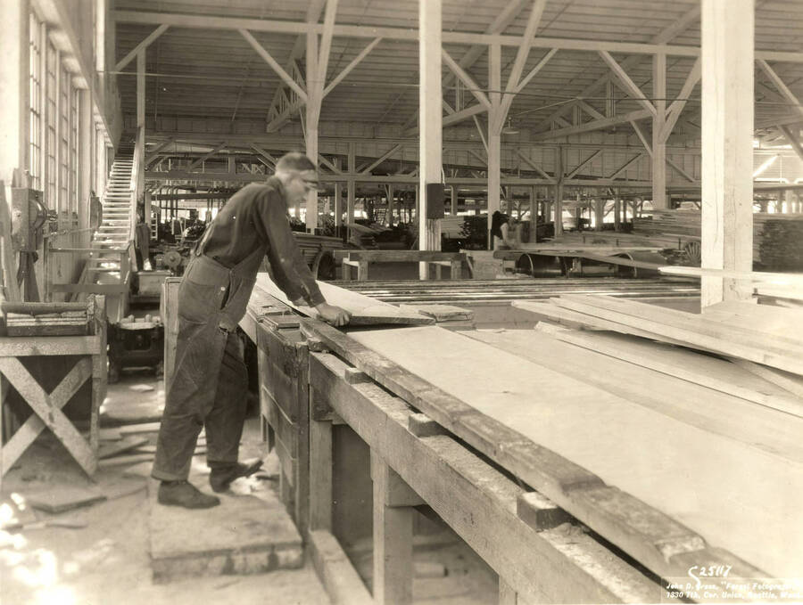 A man works with a plank of wood at the Rutledge Mill. The description on the back of the photograph says 'trimmer man who regards lumber for checks, etc. using an undercut jump saw.' On the photograph is the name of the photographer: 'John D. Cress, 'Forest Fotografer' 1330 7th Cor. Union, Seattle Wash'