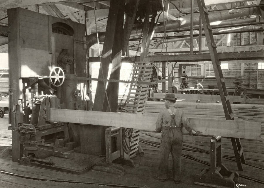 A man feeds a board of wood through a piece of mill machinery. Written on the back of the photograph 'Two McDonough and one Mershon Band Resaws in the Remanufacturing Building