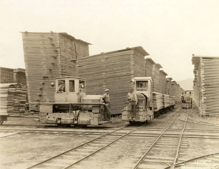 three of the Rutledge Mill lumber yard engines. Two in the foreground while one in the background. Six men control these three trains. Behind them are stacks of planks of lumber. Written on the back of the photograph: 'One of the three long alleys (No. 5) from center north, with three of the gasoline yard engines.'
