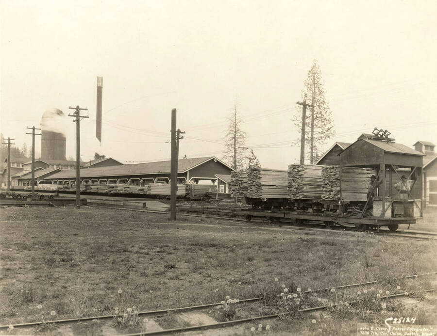 Two men ride a trolley to transfer wood from the sawmill to the lumber yard. Written on the back of the photograph: 'Trolley transfer with four cars of lumber leaving the mill sorter for the lumber yard.' On the photograph is the name of the photographer: 'John D. Cress, 'Forest Fotografer' 1330 7th Cor. Union, Seattle Wash'