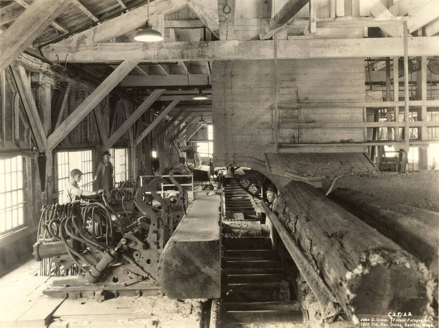 Two men work on sawmill machinery while one watches. Written on the back of the photograph: 'Martin air dogs on the left hand band mill in saw mill. On the photograph is the name of the photographer: 'John D. Cress, 'Forest Fotografer' 1330 7th Cor. Union, Seattle Wash'