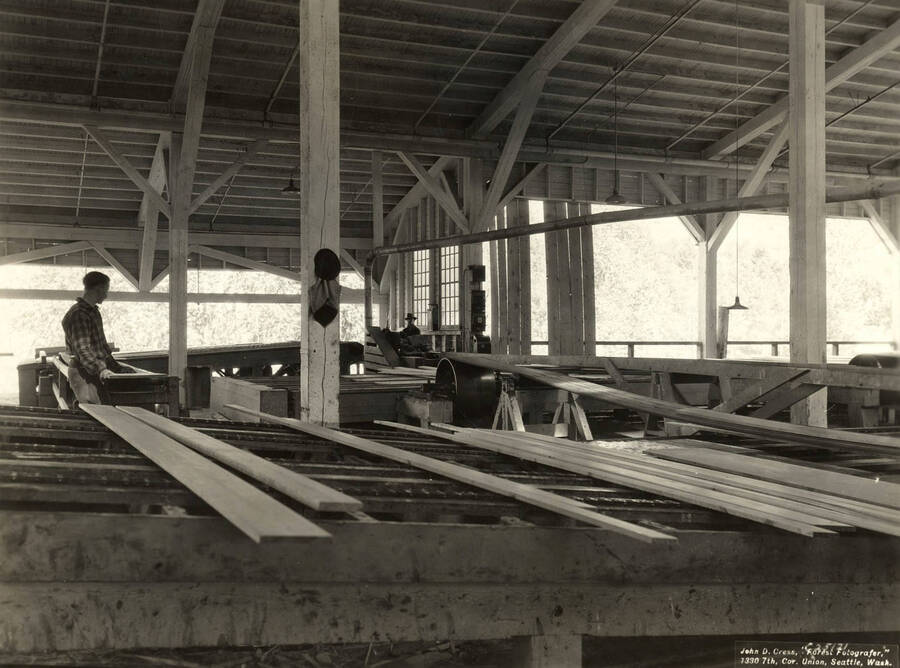 Two men work with planks of lumber. Written on the back of the photograph: 'Looking across chains and cross belts to edger.' On the photograph is the name of the photographer: 'John D. Cress, 'Forest Fotografer' 1330 7th Cor. Union, Seattle Wash'