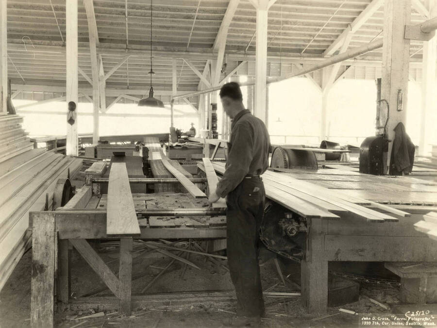 A man sends planks through a piece of machinery. Written on back of the photograph: 'Looking across transfer belts to the edger.' On the photograph is the name of the photographer: 'John D. Cress, 'Forest Fotografer' 1330 7th Cor. Union, Seattle Wash'