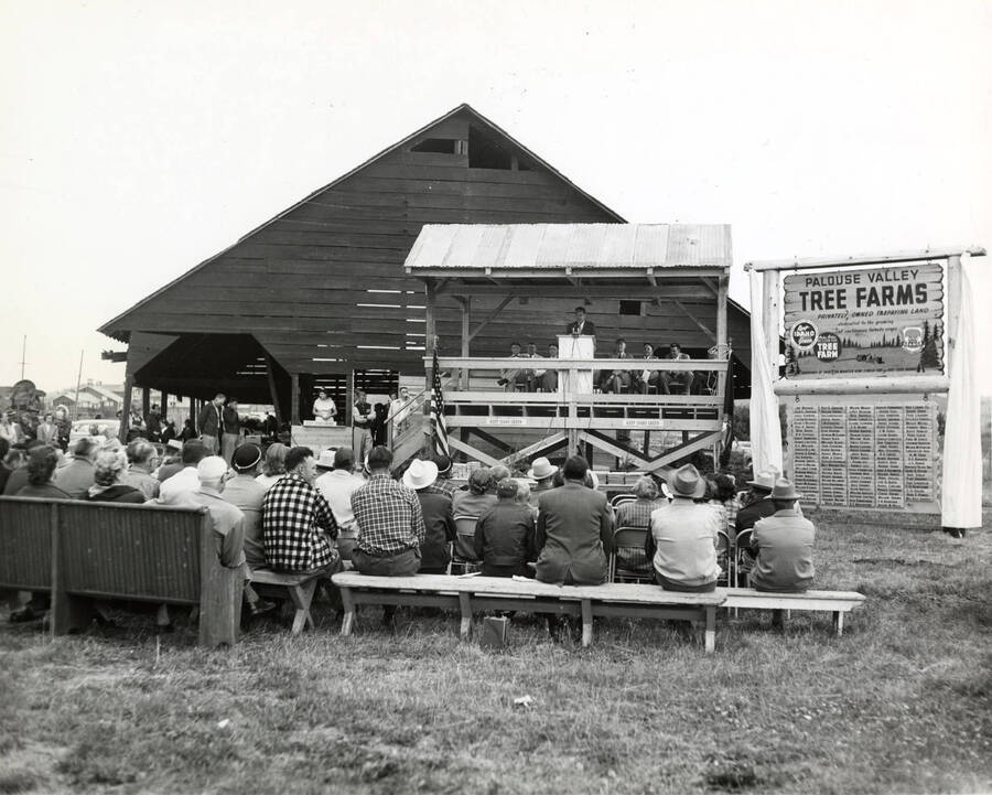 Men and women sit and listen to a speaker on Keep Idaho Green (Description taken from back of photograph). To the left of the speaker platform is a sign that reads "Palouse Valley Tree Farms."