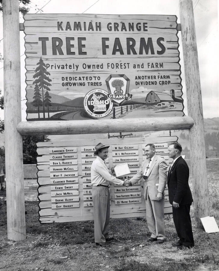 A man receives his certificate for planting trees. To his left stands two officials. They all stand underneath a sign that reads 'Kamiah Grange Tree Farms' as well as names of others who participated in the program. The description the back of the photograph reads 'Western Pine Tree Farm dedication'