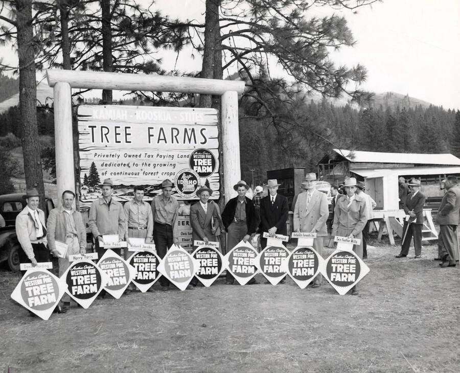Ten men receive their 'Another Certified Western Pine Tree Farm' placards and certificates as they stand under a sign that reads Kamiah, Kooskia Sites Tree Farms'. From left to right: Daniel Harding, Harry Faris, Arlie Foss, Robert Jacobs, Chris Landmark, Axel Klundt for Klundt Brothers, Ed Strehlow, Lowell Maynard, E.C. Rettig for Potlatch Forests, Inc. and Tim Waidef for Twin Feather Mills.