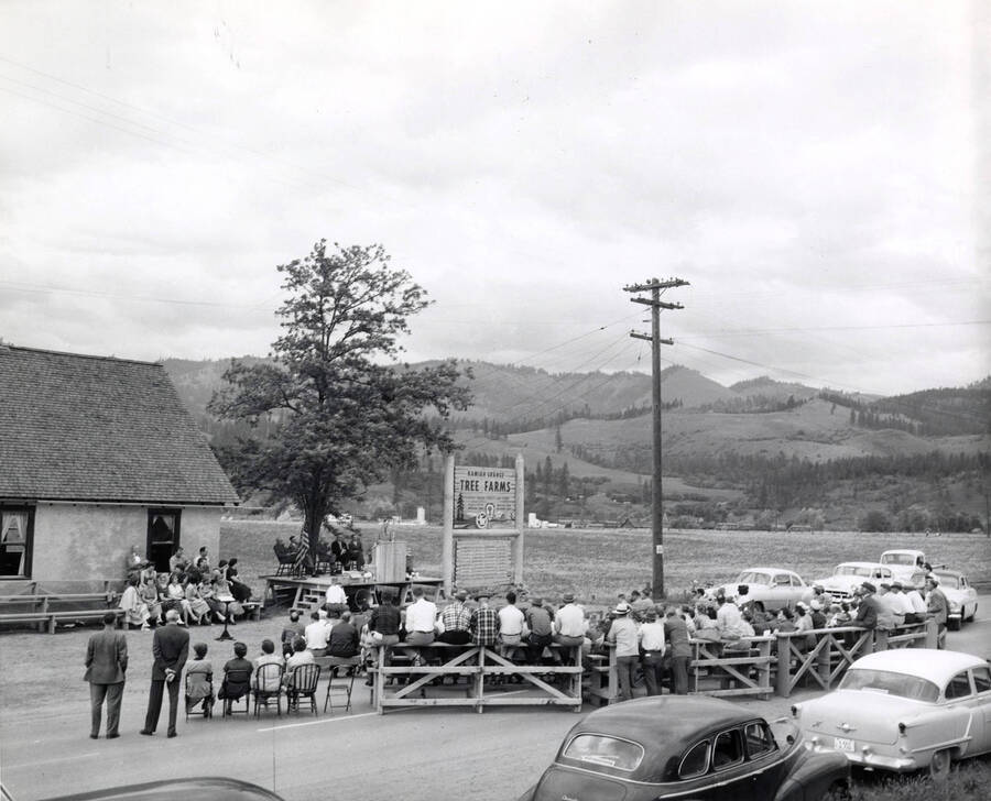 Men and women sit for a Western Pine Tree Farm Meeting (description taken from back of photograph).  To the left of the speaker platform a sign reads "Kamiah Grange Tree Farms."