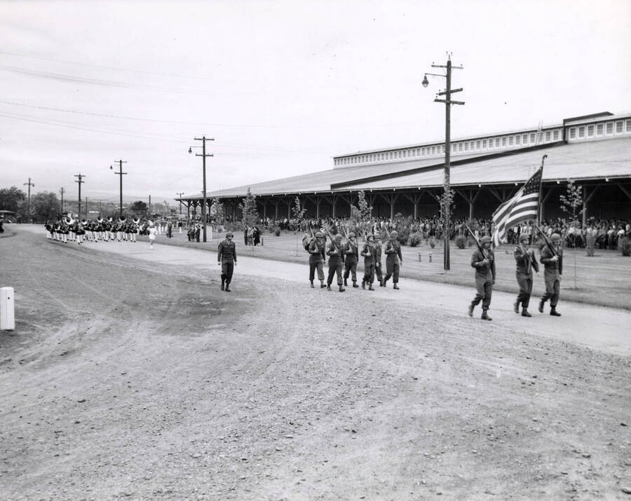 Uniformed men march down the street as part of a Memorial parade (description taken from the back of the photograph). Behind them march a band. Crowds gather underneath a building's overhang to watch.