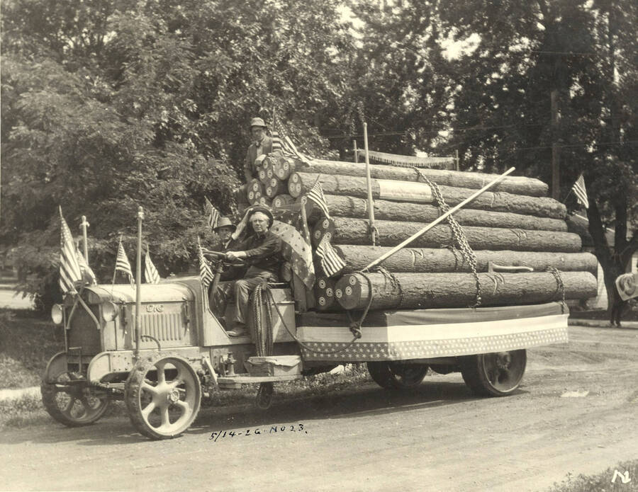 Three men ride a tractor pulling a trailer full of logs for a parade. Written on the back of the photograph is "Hard Rubber-tired truck in a local parade about 1926."