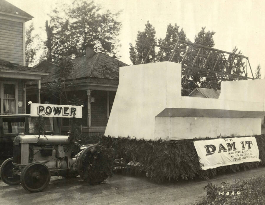 A tractor pulls a float for a parade. The theme of the float is a dam. On the side of the trailer is a sign that reads ' We Dam It' Inland Power & Light Co, Winston Brothers, Co and Fegles Construction Co'  Stamped on the back of the photograph is 'J.F. Anderson, Photo, Phone 1445-J'