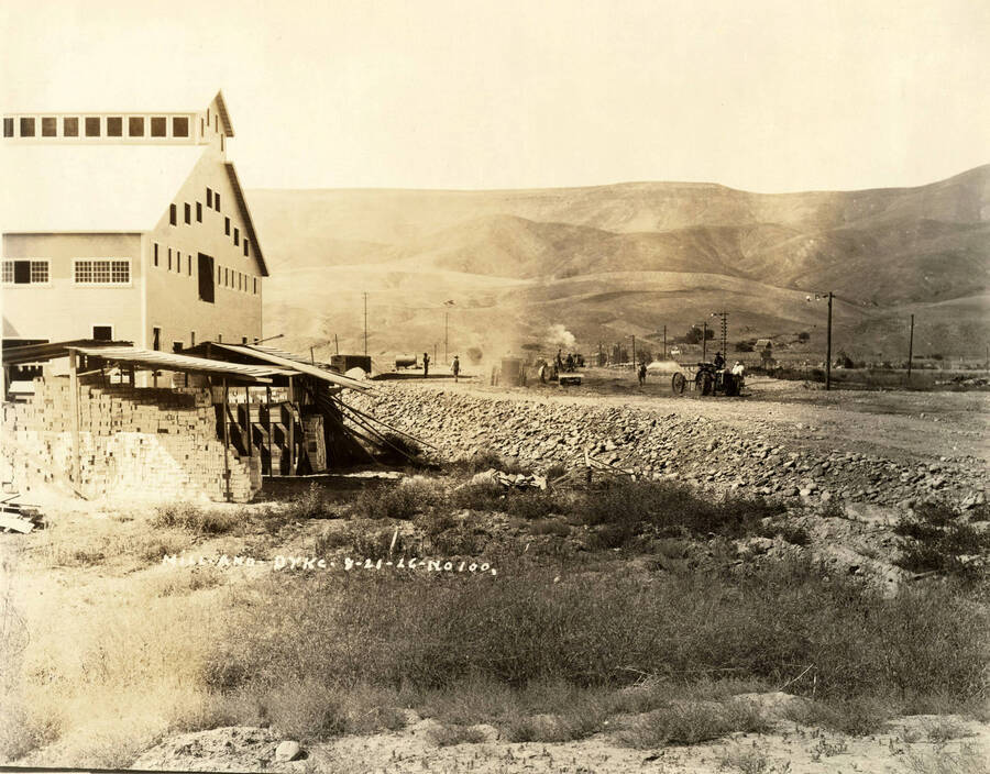 Men use tractors to construct the dike at the Lewiston Mill. On the left-hand of the photograph is the completed mill. In front of the side of the mill are piles of lumber under roofs. Written on the photograph is 'Mill and Dike 9/21/1926 No. 100'